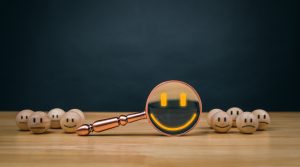 A magnifying glass, a symbol of tools to fix your online reputation, lies on a wooden table, enlarging a smiley face drawn on a small wooden sphere. Several similar spheres with smiley faces cluster nearby against a dark background.