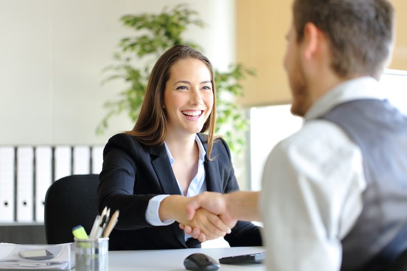 A woman in business attire smiles while shaking hands with a man across a desk, highlighting the importance of first impressions—and why your online reputation matters in today’s professional world.