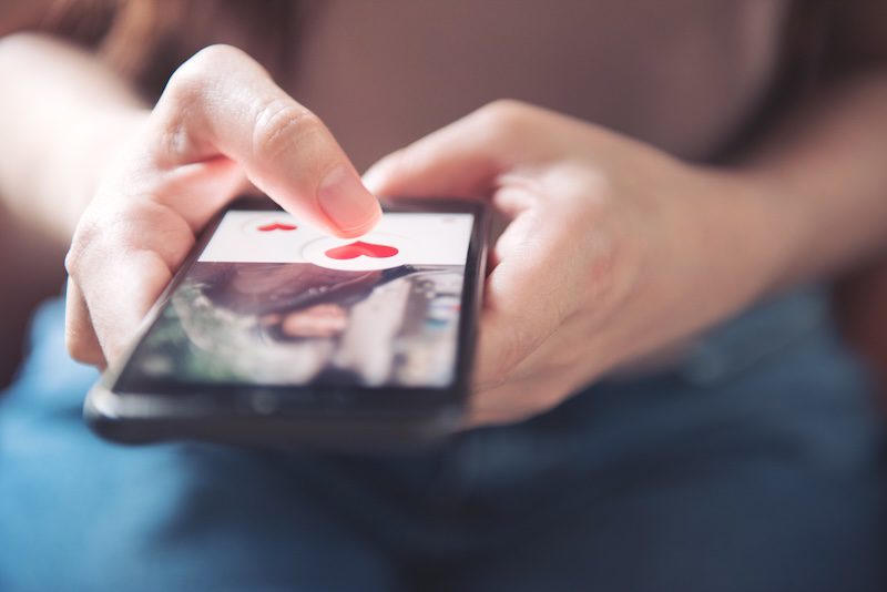 A close-up of a person’s hand using a smartphone, their finger poised to tap a heart icon—highlighting how every interaction shapes your online presence and why your online reputation matters on dating or social media apps.