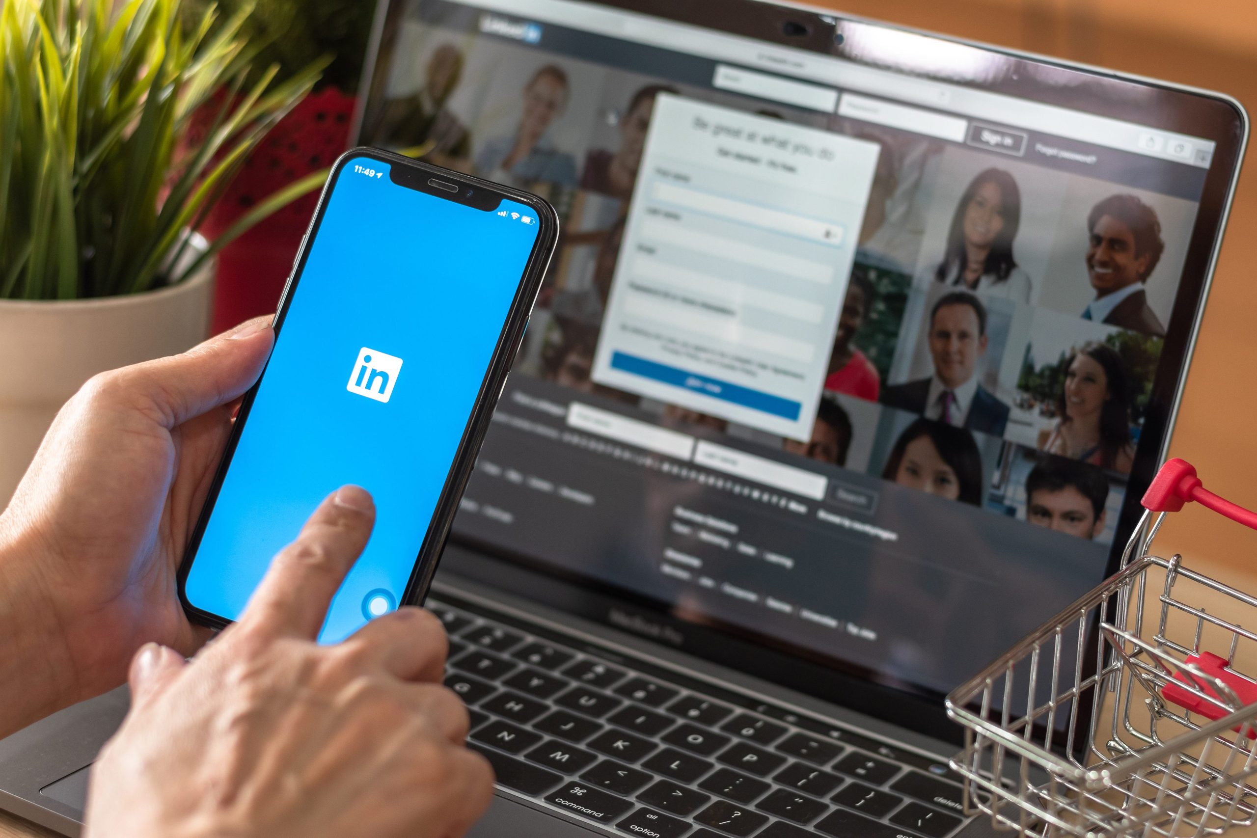 A professional at work browsing linkedin on a smartphone with a laptop in the background displaying more LinkedIn profiles, symbolizing the integration of social networking in modern business practices and illustrating How to Optimize Your LinkedIn