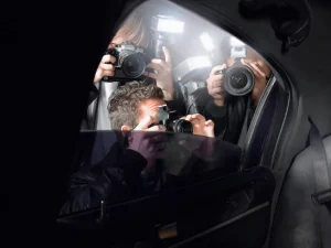 Three photographers holding cameras peering into a celebrity's car window, aiming their lenses at someone inside under dim lighting.