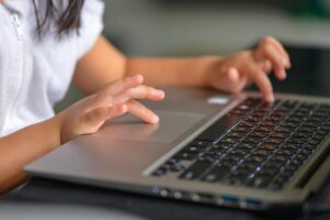 A child wearing a white shirt types on a laptop keyboard, likely taking an online IQ test, with hands and part of the arm visible. The screen is out of view, and the background is blurred.
