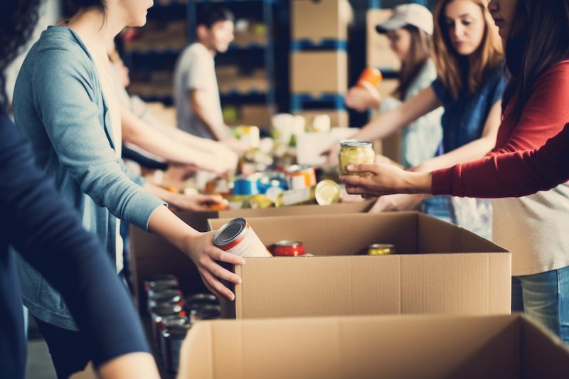 People pack food items, such as canned goods and jars, into cardboard boxes on a table in a warehouse or community center, working together as a group to support local reputation management plans.