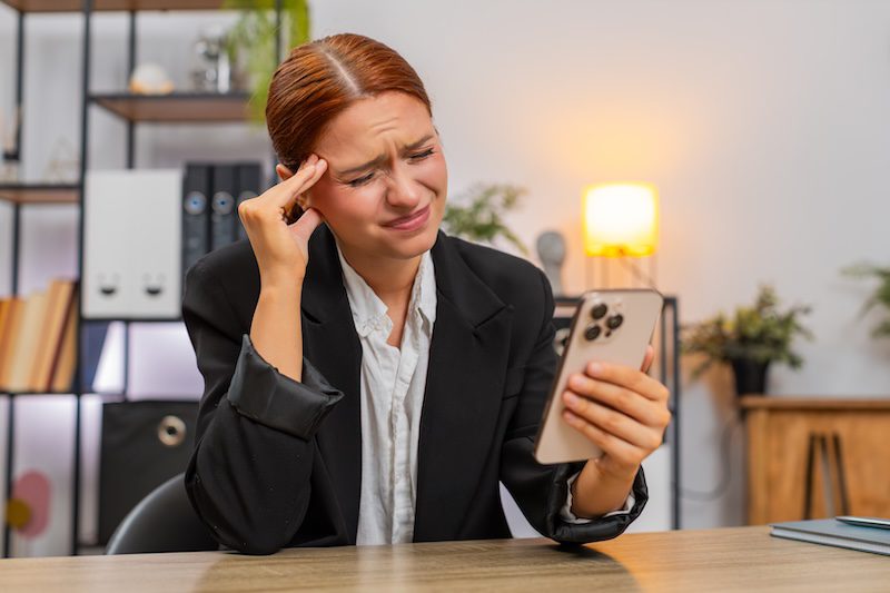 A woman sits at her desk and looks upset at a negative news article on her phone.