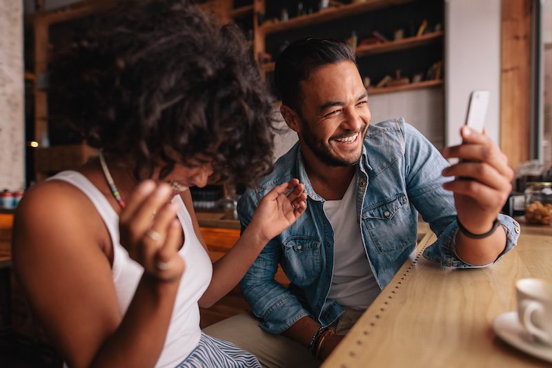 Two friends sit at a wooden table in a cafe, laughing together as one shows something funny on a smartphone—capturing a genuine moment perfect for personal branding in a relaxed, casual setting.