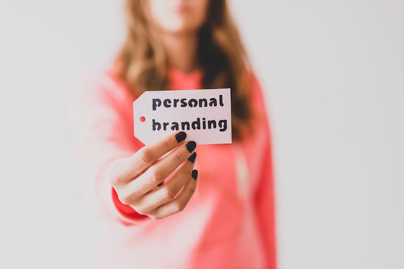 A woman in a pink sweater holds a tag labeled "personal branding," with the focus on her outstretched hand and the meaningful message.