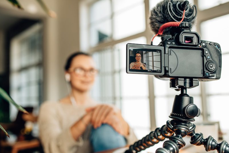 A woman wearing glasses and earbuds sits indoors, talking to a camera mounted on a tripod with a microphone, creating personal branding content. Her image appears on the flip-out screen as natural light streams through large windows behind her.