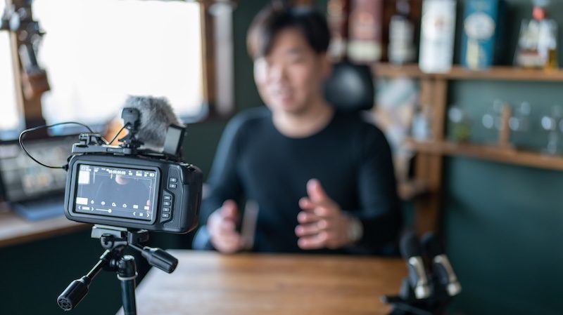 A camera on a tripod films a person at a desk, gesturing as they deliver a YouTube apology. Shelves with bottles and books in the background add to the cozy indoor setting.