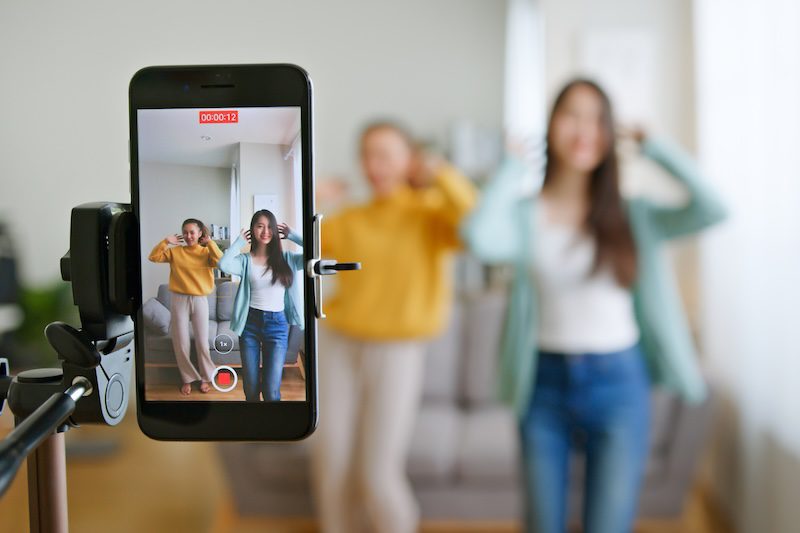 Two women pose and smile in a living room, recording a video together on a smartphone mounted on a tripod—perfecting their tiktok reputation as the phone screen captures them in focus while the real scene is blurred behind.