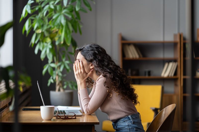 A woman with long curly hair sits at a desk with a laptop, covering her face with her hands in stress—perhaps overwhelmed while searching for the best reputation management services. A coffee mug, notebook, plant, and bookshelves fill the scene.