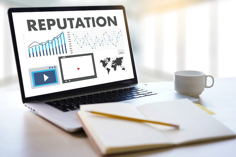 A laptop displays the word "REPUTATION" above charts, graphs, a video icon, and a world map—hinting at what is online reputation management. An open notebook, pen, and coffee cup sit on the desk in front.
