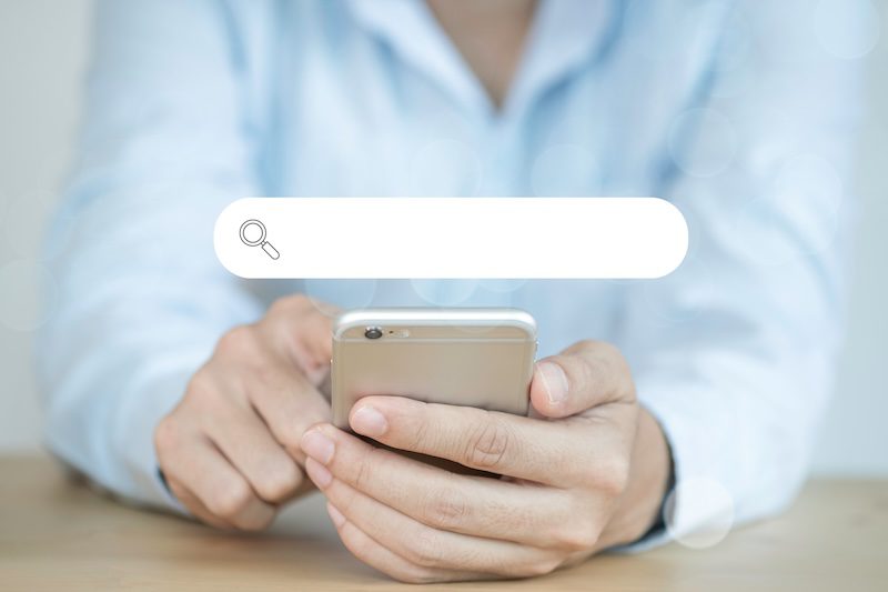 A person in a light blue shirt holds a smartphone and uses their index finger to touch the screen. A digital search bar reading "best reputation management services" with a magnifying glass icon is overlaid above their hands.