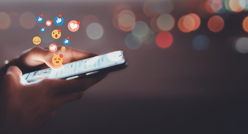 A person holds a smartphone, with social media reaction icons like hearts, thumbs up, and smiley faces floating above the screen—illustrating the power of the best reputation management services. The background is blurred with bokeh lights.