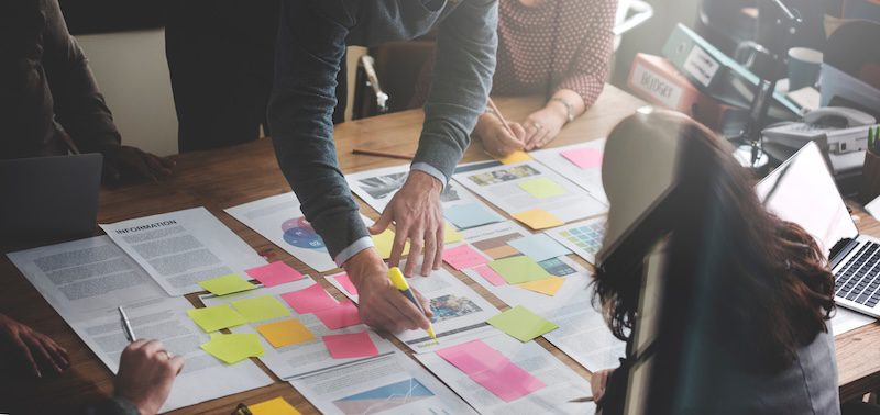 A group of people collaborate at a table covered with papers, laptops, and colorful sticky notes, discussing strategies for free online reputation management as one person highlights key points on a document.
