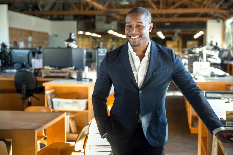 A smiling man in a navy blazer and white shirt stands confidently in a modern, open-plan office—an ideal setting to discuss what is online reputation management and its impact on today's professional environments.