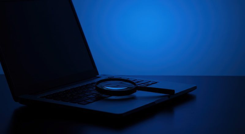 A magnifying glass rests on a laptop keyboard, bathed in blue light, evoking the atmosphere of an investigation—perfect for exploring how to deep search someone online.