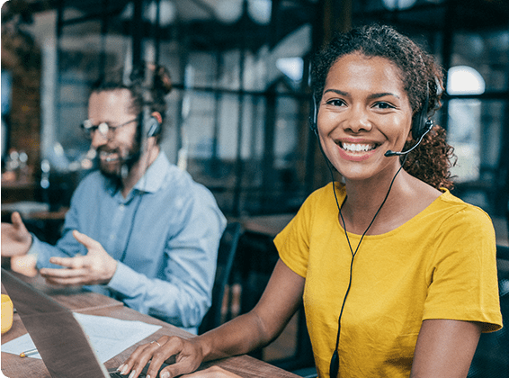 A cheerful customer service representative wearing a headset smiles at the camera, with a male colleague also wearing a headset seen talking in the background in a modern office.