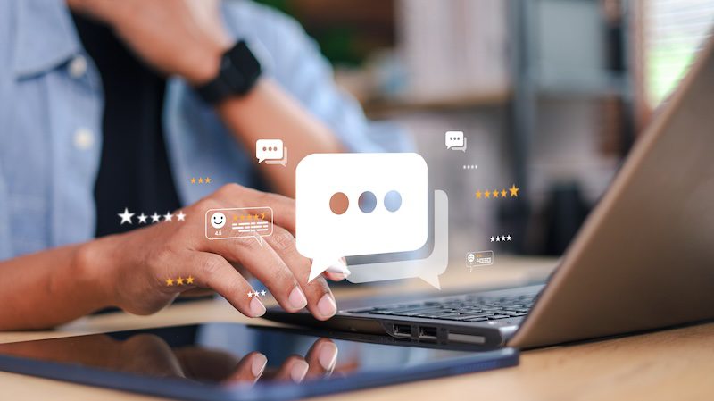 A person using a laptop with a tablet nearby, touching the keyboard. Floating digital chat and message icons are overlaid, hinting at online communication or searching for tips like how to delete a Glassdoor review.