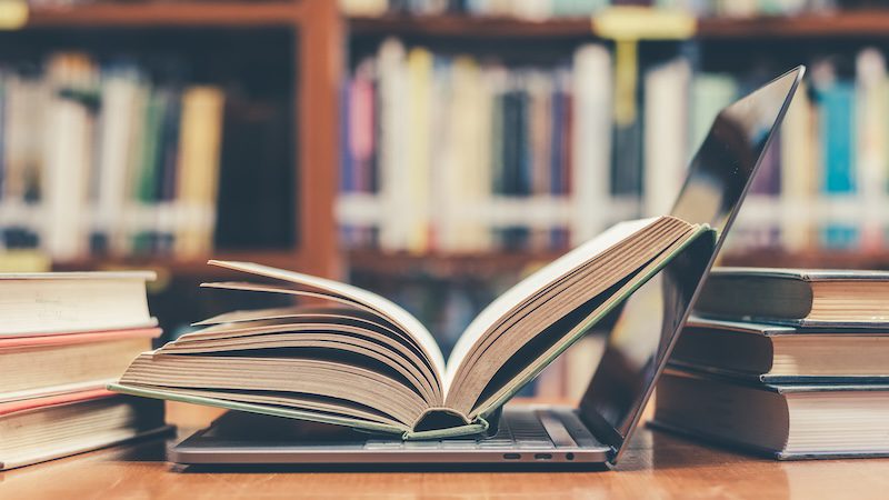 An open book rests on a laptop keyboard, surrounded by stacks of books, with blurred bookshelves behind—evoking a study or research setting and prompting the question: is Wikipedia reliable for academic work?.