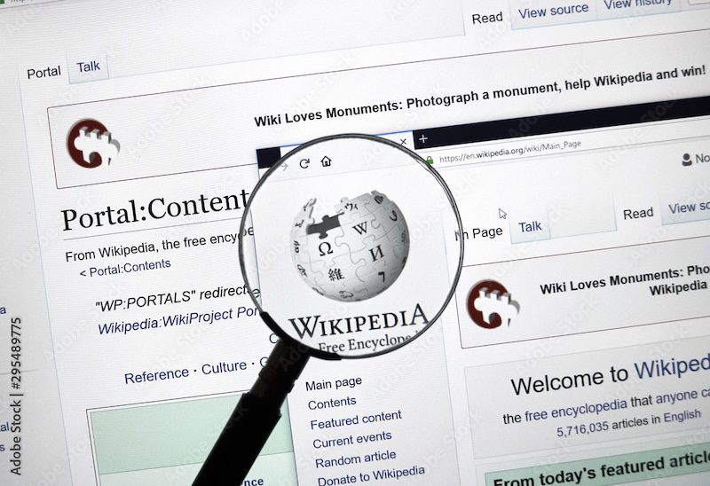 A magnifying glass zooms in on the Wikipedia logo displayed on a computer screen, with multiple Wikipedia web pages open in the background, prompting the question: is Wikipedia reliable for accurate information?.