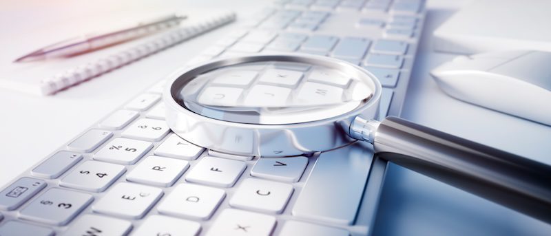 A magnifying glass rests on a white computer keyboard, with a notebook and mouse nearby, suggesting investigation or searching—perhaps even questioning, is Wikipedia reliable for the information found?.