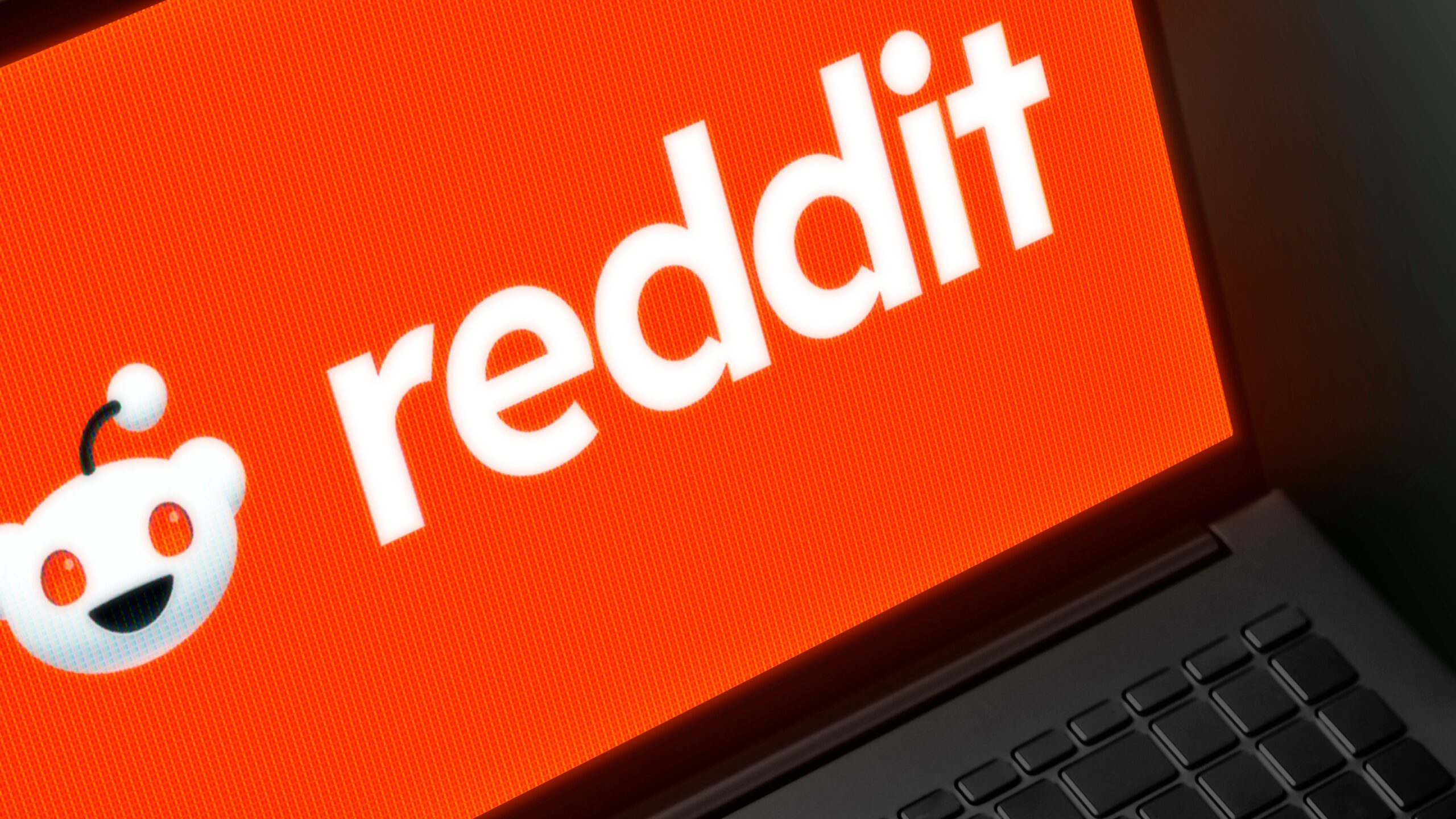 A laptop screen displays the Reddit logo in white text with the Reddit alien mascot on a bright orange background. Part of the laptop keyboard is visible at the bottom of the image.