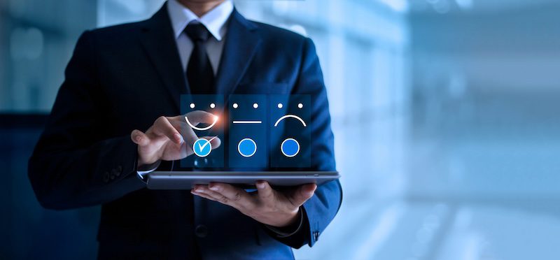 A person in a suit uses a tablet with floating icons displaying happy, neutral, and sad faces, representing a digital satisfaction or feedback survey to help fix negative online reviews.