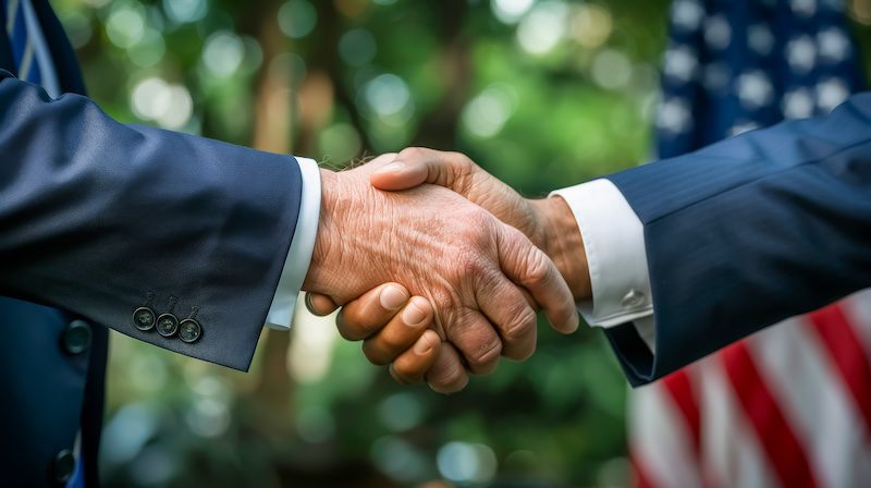 Two people in suits shake hands outdoors, with a blurred U.S. flag visible in the background.
