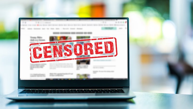 A laptop screen displays a blurred news website with the word "CENSORED" stamped in bold red letters across the screen, symbolizing restricted or blocked online content.
