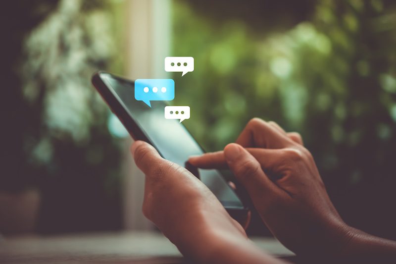 Close-up of a person’s hands holding a smartphone and texting, with digital chat bubbles appearing above the phone. The background is blurred and green, suggesting an outdoor or natural setting.