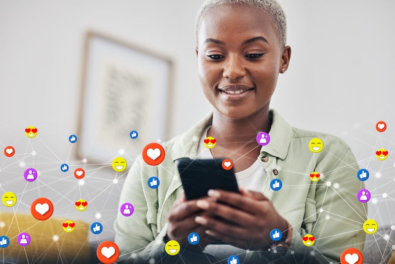 A woman smiling while using her smartphone, surrounded by floating social media reaction icons such as hearts, thumbs up, and smiley faces, representing online engagement and connectivity.