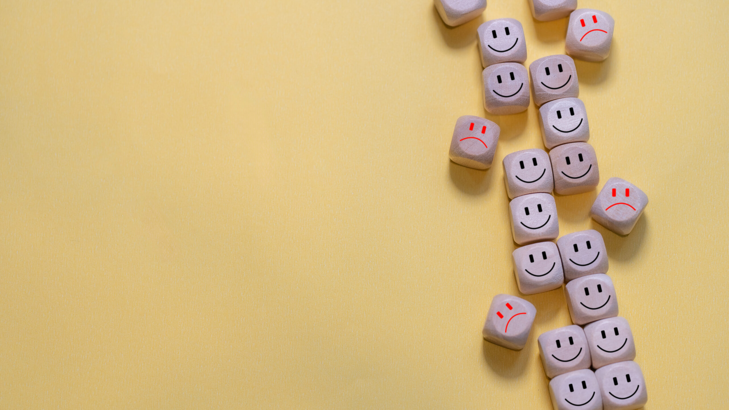 Several wooden cubes with black smiley faces and a few with red sad faces are arranged vertically on a yellow background, leaving empty space on the left side.