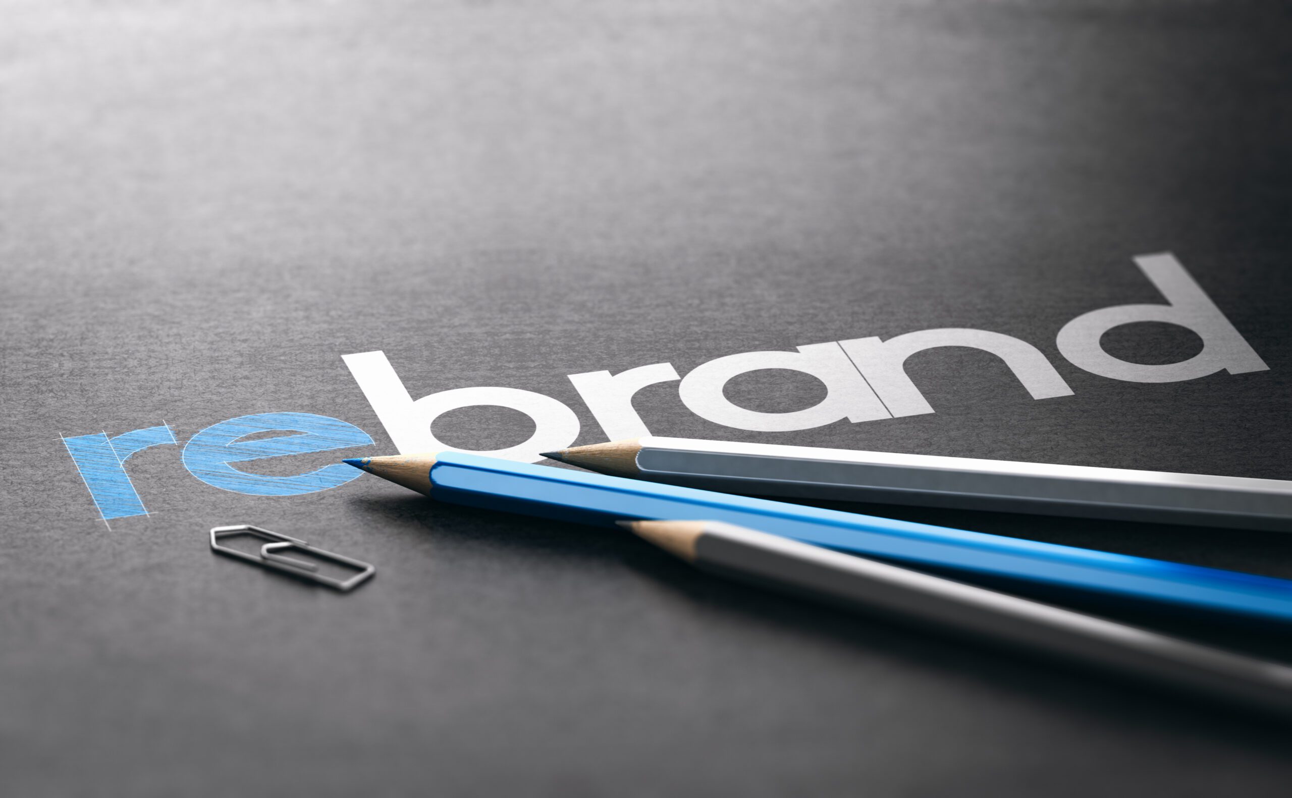 The word "rebrand" is printed on dark paper, with "re" in blue and "brand" in white. Three pencils and a paperclip are placed on top of the paper, near the text.