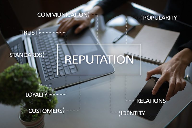 A person works at a glass desk with a laptop, notepad, phone, and plant. Words like "reputation," "trust," and "loyalty" connect by lines, illustrating the importance of Glassdoor reputation management in today’s workplace environment.